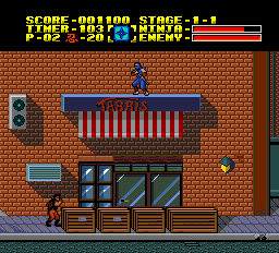 A ninja character in blue stands centered on a brick building facade during daytime. The HUD displays score, nation, stage 1-1, time remaining, and power/enemy health bars in yellow and orange text at the top. The scene shows red brick walls, wooden shutters, a blue window, and ground-level wooden crates. A small red enemy figure appears in the lower-left corner. The sprite resolution is characteristic of 16-bit PCE graphics with a limited but vibrant color palette.