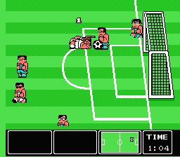 A soccer field viewed from above with bright green grass and white line markings. Multiple red and blue uniformed player sprites are positioned across the field, with one player actively controlling the ball near center. A goal net with grid pattern occupies the right side. The bottom of the screen displays a black status bar with UI elements, including a timer showing remaining match time. The sprite art uses 8-bit pixel resolution typical of NES graphics.