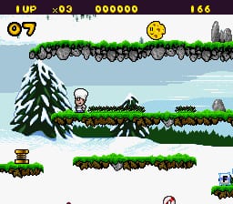 A pixel-art platformer screen displays a character standing on a grassy ledge in the center-left area. The background shows a snowy mountain landscape with evergreen trees, a light blue sky, and white clouds. The HUD at the top shows player lives (⊙7), a score counter (000000), and a level indicator (166). Green grass platforms with white snow patches form the level geometry, while a small enemy sprite appears on the lower right. The art style uses 16-bit SNES-era sprite resolution with a muted winter color palette of greens, whites, and blues.