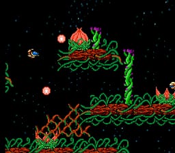 A space-themed shooter level displays a small pixelated spacecraft at the bottom center with a pinkish hue, positioned against a black starfield background. Green and orange vine-like or tentacle-like obstacles form horizontal platforms and barriers across the middle and upper portions of the screen. Two pinkish spherical projectiles are visible in mid-flight. The visual style uses low-resolution sprites typical of early NES games, with a limited color palette dominated by black, green, orange, and pink tones.