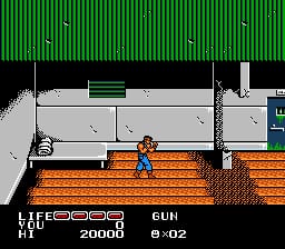 A blue-clothed player character stands on an orange brick platform in the center of the screen, with gray concrete structures and green corrugated metal visible above. The HUD displays "LIFE 2000" and "GUN 9-2" in white text at the bottom left. Brown wooden flooring stretches across the lower portion, and gray walls frame the scene on both sides. The sprite-based graphics use a limited NES color palette with black, orange, gray, and green as dominant colors.