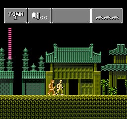 A horizontal side-scrolling level displays a character in yellow and white clothing standing on a brick platform in the lower-center area. The background features a dense arrangement of green pixel-art buildings, pagodas, and structures in varying shades of green and brown. A tall pink or magenta vertical structure appears on the far left. The HUD at the top shows a score panel with "TOWN" text, a life meter, and a pause icon. Mountains or clouds appear in the top-right corner. The environment uses a palette of greens, browns, blacks, and grays typical of NES-era sprite graphics.