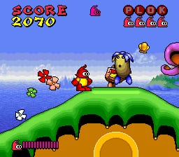 A colorful SNES-era platformer level displays a curved green hill landscape with a blue sky background. The protagonist, a red character, stands center-left on the green terrain alongside smaller companion characters. A score readout reading "2070" appears in red text at the top-left, with a power/health bar showing four filled segments in the top-right corner. Small enemies and collectibles are scattered across the rolling hills, and a pink object appears at the far right edge of the screen. The art style features chunky pixel sprites typical of early-1990s 16-bit graphics with bright, saturated colors.