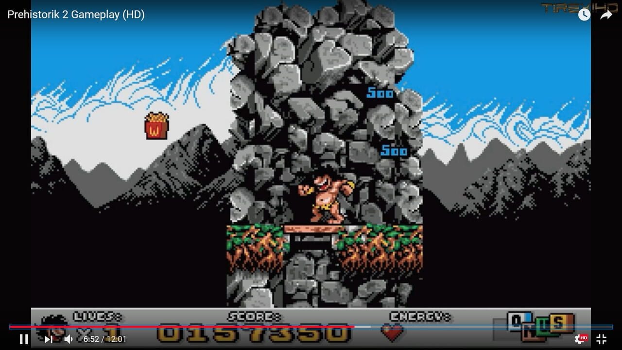 A caveman character stands on a wooden platform inside a large rocky tower structure against a mountain landscape with blue sky. Two blue treasure chest icons marked "500" float above. The bottom UI displays "LIVES", "SCORE", and "ENERGY" bars in gray. The scene uses 16-bit pixel art with parallax mountain backgrounds in shades of gray and black, typical of early 1990s DOS platformer graphics.