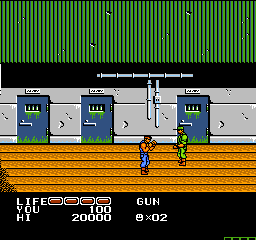 A blue-clad prisoner character stands on a wooden platform in the center of the screen, flanked by two gray industrial structures with green-lit windows on either side. The background shows a repeating green vertical stripe pattern. At the bottom left, a HUD displays "LIFE 3" and "20000" score, while text on the right shows "GUN 9-02". The sprite-based graphics use a limited NES color palette of blues, grays, yellows, and greens, typical of early 1989 NES-era pixel art.