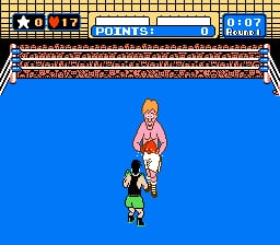 A boxing ring viewed from above shows a player character in green shorts and a smaller opponent with orange hair in a pink outfit in the center of the blue canvas. The ring is bordered by brown ropes and wooden frame details. The top of the screen displays a score panel with round and match information in red and blue sections, along with numerical values. The sprite graphics are rendered in low-resolution pixels typical of NES hardware, with a yellow and brown checkered pattern visible at the very top edge of the screen.