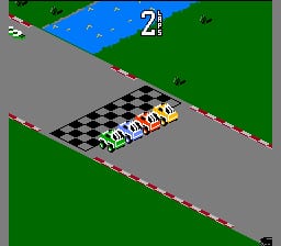 A top-down view of a isometric-perspective racing game shows a yellow and black checkered car positioned on a gray road with a checkered finish line visible. The car is surrounded by green grass terrain and gray asphalt sections arranged diagonally across the screen. A blue water area appears in the upper portion, and the number "2" is displayed in the top-right corner. The sprite-based graphics feature a low resolution typical of NES-era games, with bold color blocks defining the landscape elements.