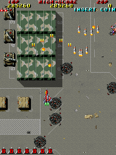 A top-down vertical scrolling shoot-em-up displays a military base layout with a large rectangular building containing a green checkerboard pattern in the center. A player-controlled aircraft appears in the upper-left area. Multiple enemy formations, including yellow and orange projectiles, scatter across the gray ground. Score and weapon indicators line the top of the screen in red text. A red ammunition counter spans the bottom. Small sprite-based explosions and debris dots the battlefield. The 16-bit sprite graphics use a muted brown and gray color palette.