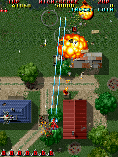A top-down vertical scrolling shooter displays a military compound with a large red rectangular building at center, surrounded by green grass and concrete pathways. A player aircraft fires green projectiles upward toward an enemy helicopter engulfed in orange explosion effects. The HUD shows score readout at top left, a minimap in the lower left corner, and a row of weapon/item icons along the bottom. Parallax layering is visible with green terrain, brown ground detail, and scattered foliage elements. The sprite resolution and color palette are characteristic of early-1990s arcade hardware.