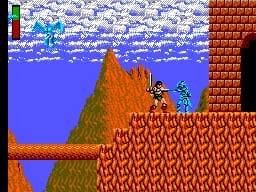 A sword-wielding warrior character stands on a red brick platform in the center-left of the screen. The background shows a blue sky with white clouds at the top, transitioning to green hillsides and orange-brown rocky terrain. Red brick walls and platforms form the level geometry, with a green grassy slope visible on the left side. The sprite uses a low-resolution pixel art style typical of early 1980s arcade ports, with the character rendered in multiple colors against the detailed brick pattern background. A narrow life bar appears in the upper-left corner.