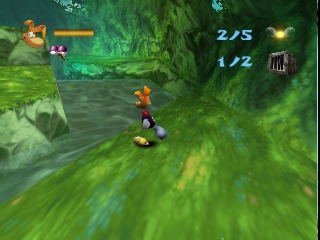 Rayman, a limbless character with orange skin, stands in the center of a vibrant green cavern with flowing water. The camera follows from a behind-the-character perspective as he faces a yellow enemy to the left. The HUD displays a score of 2/5 in the upper right corner and a health/lives indicator showing 1/2. The environment features lush moss-covered rock formations and aquamarine water with parallax scrolling effects characteristic of the N64 platformer's 3D rendering style.