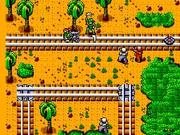 A top-down isometric view of a sandy desert level with brown stone brick walls forming pathways. Green trees and cacti are scattered across the terrain. A player character stands in the lower-left section of the screen near a vertical brick corridor. The upper portion displays a horizontal brick pathway with a vehicle and additional terrain. The color palette consists of bright yellows and oranges for sand, green vegetation, and tan/brown for structures. Sprite-based graphics with visible pixelation characteristic of SMS-era visuals.