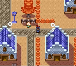 A player character stands in the center of a sandy village area flanked by two blue buildings with pointed roofs. Red spherical structures form a diagonal pattern across the upper portion of the screen. The environment uses a tan and brown color palette with a decorative tiled pattern along the top edge. A UI panel appears in the upper right corner displaying what appears to be game status information. The sprite-based graphics show 16-bit SNES-era pixel resolution with distinct architectural designs and layered environmental depth.