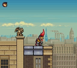 A rodent character stands on a stone platform in the center foreground, wearing red clothing. To the left sits a bronze statue on a pedestal. The background shows a detailed cityscape with tall buildings, Big Ben-style clock tower, and a bright blue sky with white clouds rendered in parallax layers. The scene uses 16-bit sprite graphics with a tan and brown stone platform in the mid-ground and pixelated architecture in the distance.