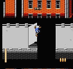 A rollerskater character in blue stands on a gray platform against a brick building backdrop with orange and red brickwork. The scene shows a two-level structure with a darker central gap, wooden crate-like platforms below, and three yellow cylindrical objects at ground level. The sprite uses limited colors typical of 8-bit NES graphics, with black serving as the primary background color separating the play areas.