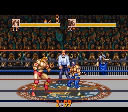 Two wrestlers face off in a boxing ring during a match. The wrestler on the left wears orange trunks, while the opponent on the right wears blue. A large ornate circular mat design fills the center of the ring floor. Health bars for both fighters appear at the top of the screen in yellow and blue. A dense crowd of spectators fills the background stands. The sprite-based graphics display the characteristic 16-bit SNES visual style with a reddish-brown color palette dominating the arena setting.