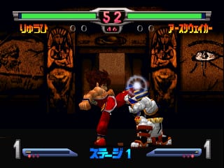 A fighting game battle screen shows two characters in combat stance on a stage with brown stone architecture and two large carved face pillars flanking the sides. The character on the left wears red clothing and appears to be executing an attack with a purple energy effect, while the opponent on the right is dressed in white and brown armor. Health bars display at the bottom corners in blue and red, with character names in Japanese text. The top center shows a tiger emblem in the HUD frame, with score counters showing zeros on both sides. The sprite-based graphics use a warm brown and orange color palette for the stage environment.