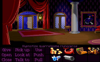 A richly decorated interior scene with red velvet curtains, ornate blue and gold doors, a gray stone column, and a framed portrait on the right wall. The floor is wooden with circular markings. At the bottom, a command interface displays verb buttons (Give, Pick, Open, Close) in purple text on the left, with a gold monkey head icon and orange text in the center, and action buttons (Look at, Push, Talk to, Pull) on the right. The pixel art uses a warm brown and red color palette typical of early 1990s adventure games.