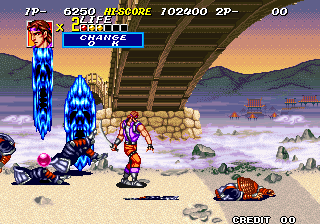 A horizontally-scrolling arcade action scene displays two player characters engaged in combat on a beach landscape. Blue flame-like effects emanate from the left side where enemies are grouped. The top HUD shows score (6250), stage difficulty indicator, and player lives (2P- 00). Sandy terrain with scattered rocks fills the foreground, while mountains and a wooden pier structure frame the background under a sunset sky. Sprite-based pixel art renders all elements across the wide arcade screen with parallax layering visible in the distant scenery.
