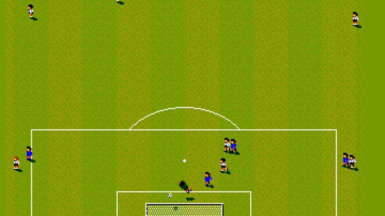 A pixelated overhead view of a soccer match in progress on a grass field with white boundary lines and a center circle. Two teams in contrasting uniforms (white/red and blue) are positioned across the field with players scattered in various formations. A goalkeeper stands near the goal area at the bottom, and a small white ball is visible in the center. The goal net is shown with a dotted pattern, and several player sprites occupy the midfield and defensive positions. Two additional players appear at the top edges of the field, with one small black sprite visible above the field.