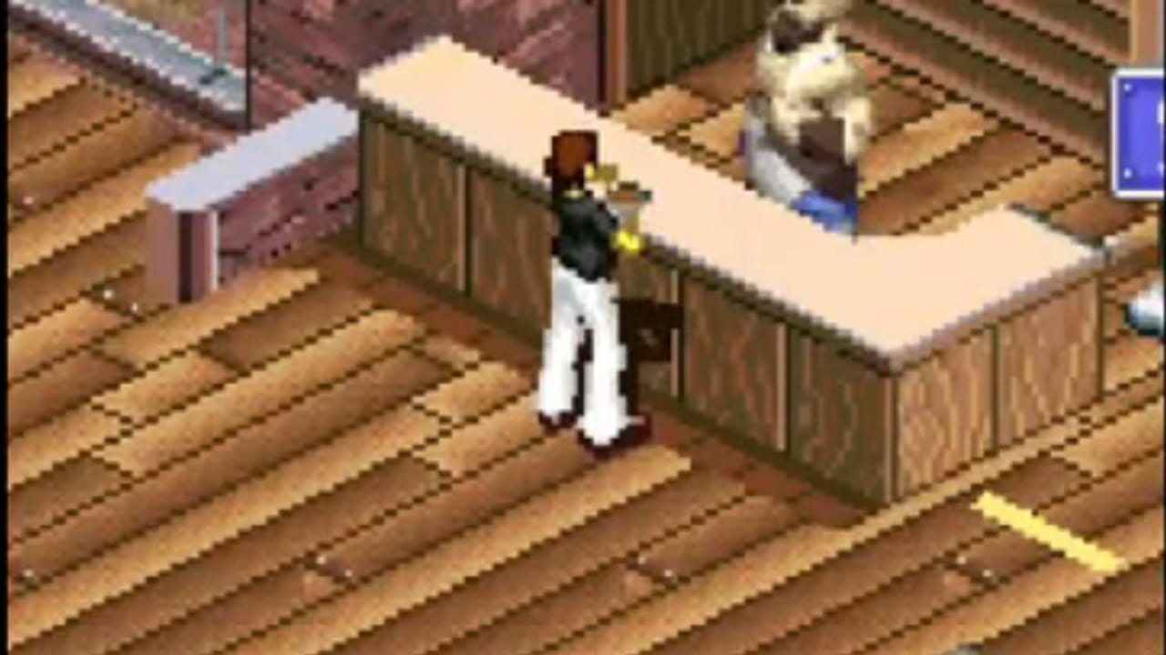 An isometric view of a residential interior shows a character with a brown head and black torso wearing white pants standing on a wooden floor. A cream-colored counter with brown wooden cabinets is positioned to the right, with a white cat resting on top. Additional furniture including a bed frame and brown walls frame the space. A blue UI element appears in the top-right corner. The pixel art uses a warm color palette with detailed wood texturing on the floor and architectural elements.