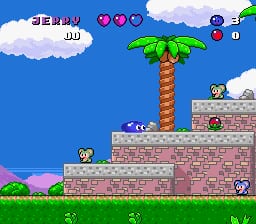 A brick platform level with a blue sky background and white clouds at the top. A brown palm tree with green fronds stands in the center-right area. The player character, a small blue ball, is positioned on a tan brick platform in the lower portion. A red brick wall structure forms the main level geometry on the right side. The top-left corner displays the game's HUD with a score counter and health indicators shown as colored circles. Additional UI elements and score displays appear in the top-right corner against the bright blue sky background.
