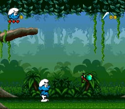 A blue Smurf character stands on grass in the foreground of a jungle level, facing right toward a red enemy creature. Hanging vines and dense green foliage fill the mid-ground, with a wooden branch extending across the upper portion of the screen. The background shows layered green vegetation with blue sky visible at the top. Two icons appear in the upper corners—one displaying a Smurf head and one showing a mushroom—likely representing health and collectibles. The sprite-based graphics use a bright 16-bit color palette typical of SNES platformers.