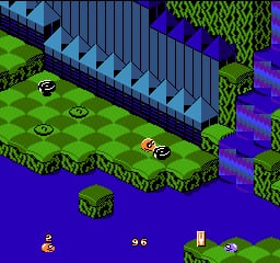 An isometric 8-bit level displays two yellow snake characters on a bright green checkered ground surrounded by blue water. A long blue fence structure runs diagonally across the upper portion. Green trees and purple bushes populate the terrain edges. The top-left shows a score display reading "26", with additional small yellow sprites visible in the lower-left corner. The scene uses a limited NES color palette of bright greens, blues, yellows, and purples with visible pixelation typical of early 1990s cartridge graphics.