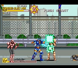 A fighting game battle scene displays three characters on a street stage with a brick wall background. On the left stands a female fighter in red and white outfit, center shows a blue-suited male character mid-combat, and right displays a cyan-colored opponent. The HUD shows round information (15) at top left, a health bar system across the upper portion, and stage/character icons at bottom. Pixel art sprites are rendered in 16-bit resolution against a layered urban background with green bushes and stone wall texture. The interface uses gold and gray UI elements typical of early-90s fighting games.