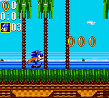 Sonic runs rightward across a grassy platform in a side-scrolling level with a cyan sky and brown ground. The HUD displays score, time, and ring count in the upper left corner with colored numerical readouts. Three gold rings float above the ground ahead of Sonic. Mountains and clouds appear in the distant background. The sprite-based graphics use a limited color palette typical of Game Gear hardware, with distinct pixelation and simple environmental details.