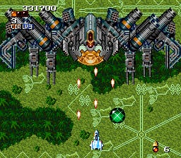 A top-down vertical scrolling shooter displays a symmetrical fortress structure with multiple turrets arranged in rows at the center of the screen. A small player-controlled white spacecraft hovers near the bottom of the playfield against a green grass background with darker forest areas on either side. Yellow projectiles trail upward from the player ship toward the enemy installation. The score display reads 325700 in the upper left corner, with additional HUD elements visible in the top-right. The overall visual style uses Mode 7-like perspective scaling common to SNES-era shooters.