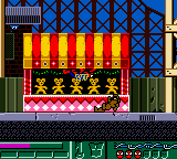 A Game Boy screenshot displays a side-scrolling platform level with a red and yellow striped building structure in the center-upper portion. Blue diagonal crosshatch pattern fills the background. Below the building sits a pink checkered band with a dark ornate section containing small colored details. A green ground platform runs across the bottom, with a black HUD bar displaying level information and a score counter at the very bottom. Sprite-based graphics rendered in the Game Boy's limited color palette of greens, reds, yellows, blues, and blacks define the scene.