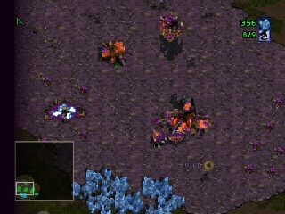 An overhead RTS view shows a dark purplish terrain with scattered units. Orange and red-colored military units cluster in the center and right side of the map. Blue units occupy the bottom-left corner near a small minimap window displaying the level layout. Teal and yellow UI indicators appear in the top-right corner. The graphics display the characteristic low-resolution sprite style of N64-era real-time strategy games, with pixelated unit models and a muted color palette typical of the StarCraft 64 conversion.