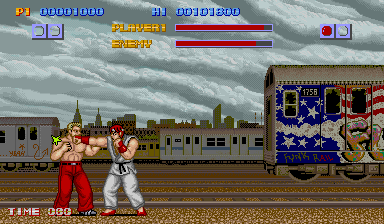 Two fighters in red and white clothing face off on a street stage with a city skyline and train in the background. The HUD displays both fighters' health bars at the top in red and blue, with time remaining and round information. The sprite-based characters and pixelated background use the characteristic 16-bit color palette and resolution typical of late-1980s arcade graphics. A decorative train car with stars and artwork is visible on the right side of the stage.