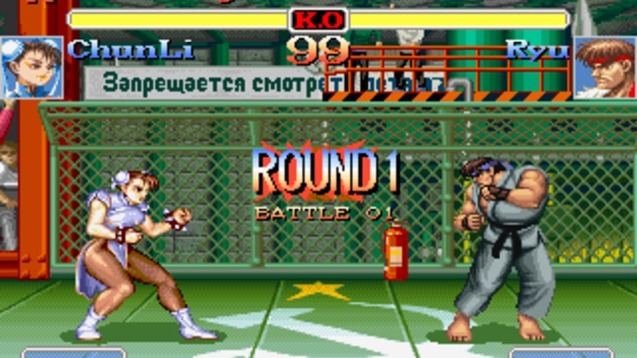 A one-on-one fighting match displays two characters facing each other on a green martial arts arena with chain-link fence backdrop. On the left stands a female fighter in a blue outfit in fighting stance; on the right is a larger male opponent in gray pants and dark top. The HUD shows character names at top left (Chunli) and right (Ryu), health bars in yellow, a large 'K.O' indicator in the center, and 'ROUND 1 BATTLE 01' text overlaying the arena. Russian Cyrillic text appears above the health bars. A red fire extinguisher sits at center arena. Character portraits occupy the upper corners.