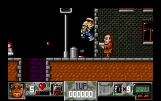 A side-scrolling platformer level displays a muscular character in a hat standing on a brown dirt platform against a brick wall backdrop. A second character in brown clothing stands to the right. The left side shows a purple-textured wall section and orange brick platform. At screen bottom runs a game interface bar with inventory slots showing items including what appears to be weapons and tools, life counter displaying 5, and a score readout showing 000000. A tall metallic cylindrical object stands center-right on the platform. The graphics use 16-bit sprite resolution with a limited color palette of browns, brick reds, purples, and blacks.