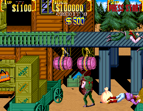 A side-scrolling scene displays a wooden building with an open doorway on the right where a character in green clothing stands. Two large purple barrels hang from chains above the building's entrance. To the left, another wooden structure with green-trimmed windows is visible. A character in purple and yellow attire appears in the lower right corner near a prone figure. The upper portion shows score counters displaying values like $1100 and $10000 against a black background, with green terrain and additional structures visible in the background. The color palette features warm browns, greens, and purples typical of early 1990s arcade sprite graphics.