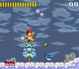 A character in a red outfit stands on a snowy landscape with a snow-covered tree in the center. The background displays a blue sky with white snowflake particles falling. A blue creature appears to the right of the tree, and gray rock formations sit at the bottom right. The top of the screen shows a yellow score bar on the left displaying health or collectibles, with "00" displayed in white text on the right. The overall color palette consists of blues, whites, and browns rendered in 16-bit sprite graphics typical of early 1990s console gaming.