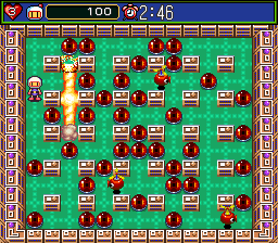 A top-down view of a bomb-themed grid arena with a white player character centered on the green playfield. Soft blocks in tan shades fill most grid spaces, with darker stone walls forming a pattern throughout. Scattered red bomb icons and power-up items dot the board. A decorative brown and tan border frames the arena edges. A status bar at the top displays the score "100" in yellow text, a timer showing "2:46", and small icon indicators on the left side. The color palette uses bright greens, reds, tans, and blues typical of SNES-era sprite graphics with visible pixel resolution.