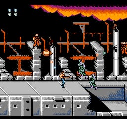 A soldier character stands on a gray platform in the lower portion of the screen, facing right with a weapon raised. Above and behind him, an orange and brown industrial structure with wooden beams, pipes, and metal platforms fills the mid-ground. A large orange and yellow explosion or fire effect dominates the top of the screen against a black sky. Two white ammunition counters appear in the upper left corner. The pixel art uses a limited color palette with orange, brown, gray, and black tones typical of NES-era graphics.