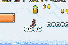 Mario stands on a platform in the center of the screen, facing right with a small outline box beneath him showing movement controls. A brown wooden platform extends from the left side. The top UI shows a score counter on the left, a timer in the center, and coin and power-up icons on the right. A yellow power-up box hangs suspended above, with more coins scattered across the upper portion. The background displays a light blue sky with white clouds. The bottom of the screen shows a row of small white blocks indicating remaining lives, rendered in the characteristic 8-bit sprite style of the Game Boy Advance.