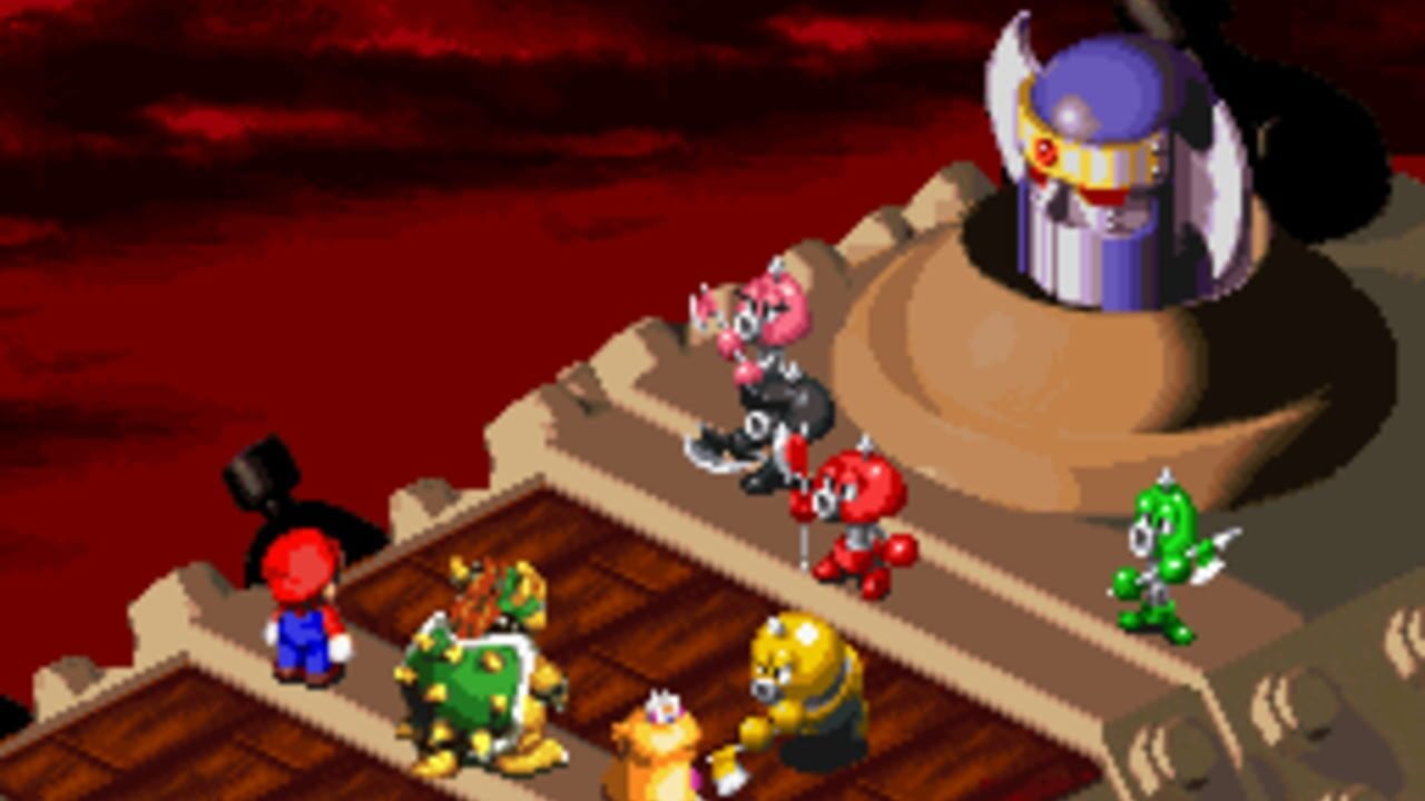 An isometric view shows Mario standing on a wooden floor to the left, with a large tan shoe or boot structure above containing multiple enemies. Pink flower-like creatures, black bomb enemies, and red round enemies are visible on the boot's surface. A green caterpillar-like enemy stands to the right. A purple and blue crowned character with white feathers appears in the upper right. The background is a dark red gradient. Sprite-based graphics with layered isometric perspective create depth in the scene.