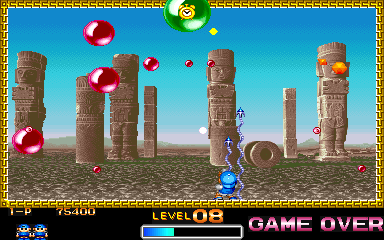 A gameplay screen displays a blue-clad character standing in the center of an ancient stone temple ruin with tall columns scattered across a desert landscape. Red and green bouncing orbs float above the terrain while smaller projectiles descend. The HUD shows "LEVEL 3" and "GAME OVER" text at the bottom, with a health bar and score information visible in the lower-left corner. A decorative gold border frames the entire screen, and the art uses 16-bit era sprite graphics with a clear blue sky background.