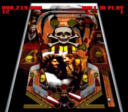 A pinball table displays a skull-faced mask character in the center playfield, surrounded by orange and yellow geometric shapes and bumpers. Red text at the top shows the score "944,219" and "BALL IN PLAY" indicator. The table layout features a dark background with contrasting bright elements typical of SNES-era pinball graphics, including flippers positioned at the bottom of the screen and various bumper targets distributed across the playing surface.