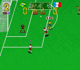 A soccer match viewed from above shows a bright green field with white boundary lines and goal areas. Two teams in contrasting colors—one in red, one in white—are positioned across the field with the ball near center. The scoreboard at the top displays numerical stats in yellow text. A goalkeeper stands in the lower-left goal area. The sprite-based graphics use a 16-bit art style with flat color blocks typical of early-1990s SNES sports games.