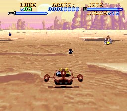 A desert landscape with a brown sand terrain dominates the screen. In the foreground center, a red vehicle with visible wheels and mechanical details moves across the sandy ground. The HUD at the top displays a life bar on the left, score and enemy count in the center, and what appears to be a timer or level indicator on the right. Two small enemy figures are visible in the mid-distance on the sand. The background shows pink and purple colored rock formations and distant terrain under a pale sky, with layered parallax scrolling creating depth. The overall pixel art style uses a tan and brown color palette typical of 16-bit SNES graphics.