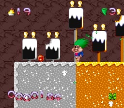 A side-scrolling level displays a troll character on gray stone ground in the lower left, with a row of lit torches and white-capped towers rising above on brown terrain. The right side shows orange crystalline blocks stacked vertically. The background features brown earth with scattered torches and multiple white-peaked structures. A UI bar at bottom displays colored coin or item icons in a row against dark background. The sprite-based art uses a limited color palette with visible pixelation typical of early-1990s SNES graphics.