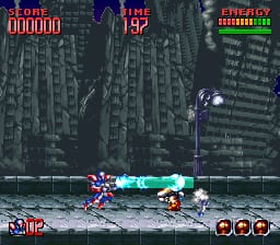 A character in cyan armor fires a weapon while standing on a gray platform in an urban environment with tall buildings in the background. The HUD displays a score of 000000 in the top-left, time remaining of 192 in the top-center, and an energy bar with remaining health indicators in the top-right. Three circular icons appear in the bottom-right corner. The pixel art features a dark color palette with detailed architectural elements and parallax layering in the background buildings.