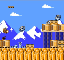 A horizontal scrolling level displays a snow-covered mountain landscape with white peaks against a bright blue sky dotted with white clouds. Golden wooden crates and boxes are stacked across the ground level, while blue shipping containers sit in the mid-section. A wooden structure with a yellow roof and blue doors occupies the right side of the screen. Two small enemy sprites with distinct patterns are visible on the platforms. The sprite-based graphics use a limited color palette typical of NES-era platformers, with clear pixelation and layered parallax scrolling creating depth.