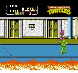 A green-skinned turtle character stands on a blue platform in the center of the screen, facing right. The top of the screen displays the Teenage Mutant Ninja Turtles II logo in orange and yellow, with a life counter and score panel in white text on the left. The background shows a street scene with white brick buildings and green doors. Below the character, an orange and red flame effect creates a hazard barrier. The sprite uses pixel art typical of NES-era side-scrolling beat-em-ups, with limited animation frames and a bright, constrained color palette.